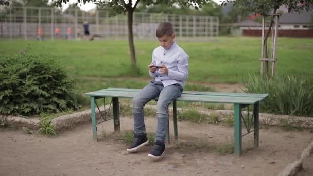 Handome niño sentado en el banco y jugar juegos en línea durante las vacaciones escolares — Vídeo de stock