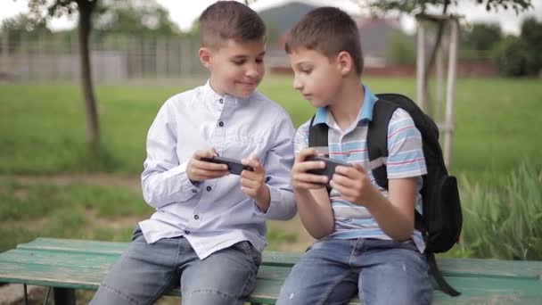 Двоє хлопчиків сидять на лавці і грають в онлайн ігри. Один хлопчик з рюкзаком. Молоді хлопці користуються своїми телефонами — стокове відео