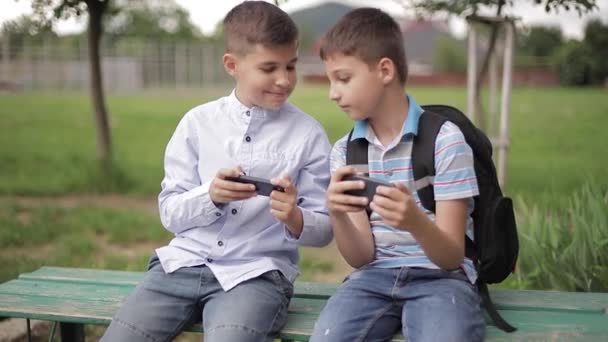 Deux garçons assis sur le banc et jouer à des jeux en ligne. Un garçon avec un sac à dos. Les jeunes garçons utilisent leur téléphone — Video