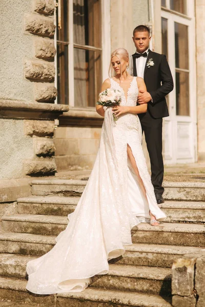 O noivo e a noiva estão nas escadas. Modelo de moda em roupas elegantes. Vestido longo branco e terno elegante — Fotografia de Stock