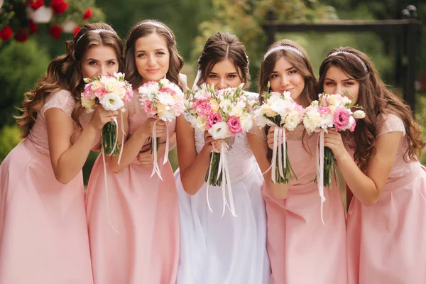 Gelukkige bruid met de bruidsmeisjes houd boeketten en veel plezier buiten. Mooi bruidsmeisje in dezelfde jurken staan door de charmante bruid in lange trouwjurk — Stockfoto