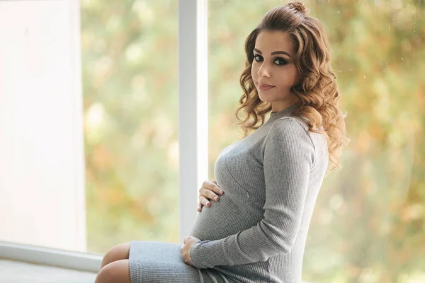 Těhotná žena seděla na parapetu u okna doma. Měla na sobě krásné těhotenské šaty — Stock fotografie