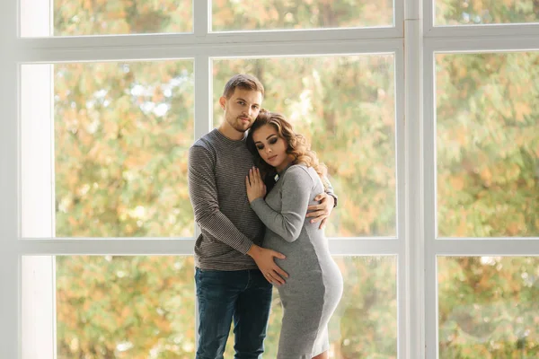 Mann umarmte seine schwangere Frau und legte seine Hand auf ihren Bauch. sie stehen am Fenster — Stockfoto
