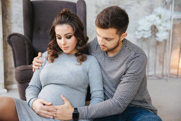 Die junge Familie sitzt neben dem Stuhl und blickt auf den Bauch. Pregman Frau mit ihrem geliebten Mann. Glückliches Paar — Stockfoto