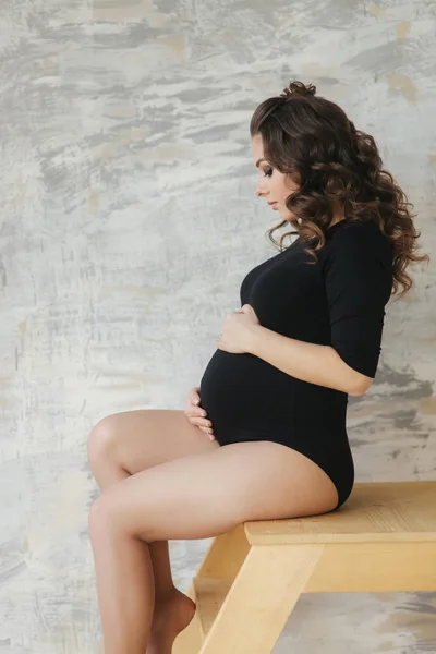 A mãe grávida senta-se nas escadas. Modelo vestido de macacão preto. Ela pôs a mão na barriga. Futura mãe feliz — Fotografia de Stock