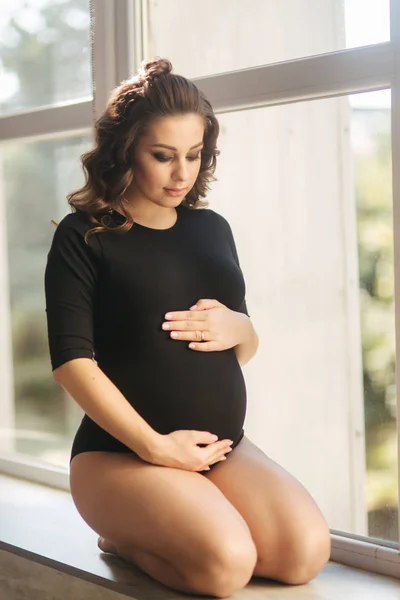La madre embarazada se sienta frente a una gran ventana. Modelo vestido con traje negro. Ella puso su mano en el vientre. Feliz futura madre — Foto de Stock