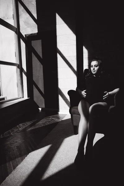 Gyönyörű terhes nő ül a széken előtt nagy ablak. Fekete-fehér fénykép — Stock Fotó