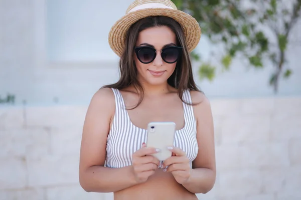 Hermosa mujer joven en traje de baño y pantalones cortos utilizan el teléfono en el trópico. Mujer con sombrero y gafas de sol — Foto de Stock