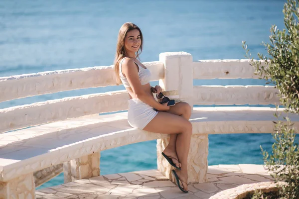 Mujer joven sentada frente al hermoso mar. Color azul del agua. Mujer en gafas de sol se sienta en el banco cerca del mar — Foto de Stock