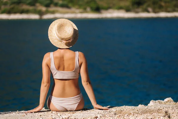 Vacker ung kvinna i hatten sitta på klippan vid den vackra lagunen. Tillbaka syn på sexig kvinna sitter vid havet. Bakgrund av blått vatten Royaltyfria Stockfoton