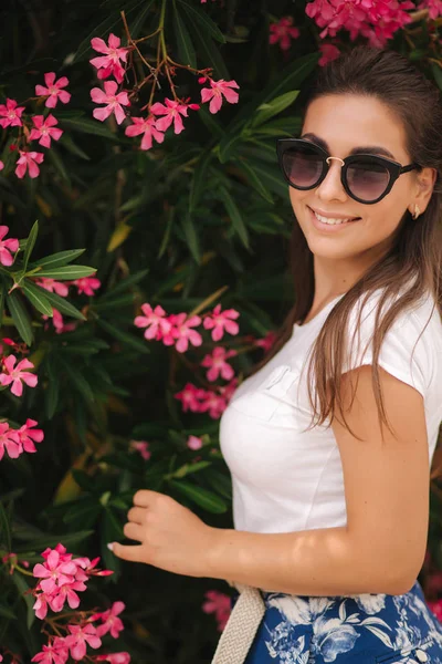 Wunderschöne Frau mit Sonnenbrille steht in wunderschönen Blumen. Porträt einer glücklich lächelnden jungen Frau — Stockfoto