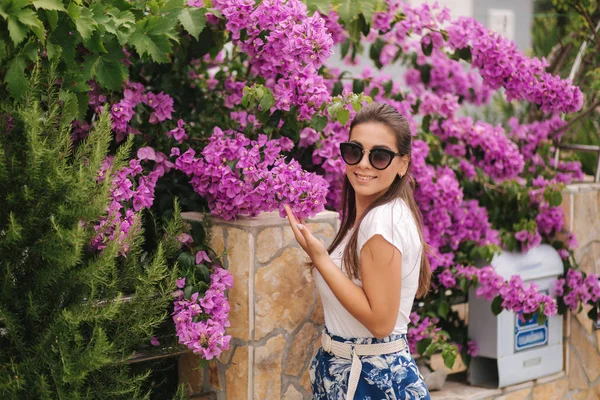 Mujer hermosa en gafas de sol de pie en hermosas flores. Retrato de la joven sonriente feliz —  Fotos de Stock