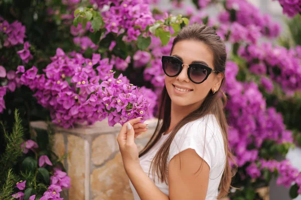 Wunderschöne Frau mit Sonnenbrille steht in wunderschönen Blumen. Porträt einer glücklich lächelnden jungen Frau — Stockfoto