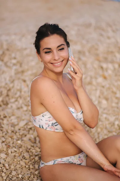 Junge und schöne Frau in Bibini sitzen am Strand und benutzen ihr Telefon. Weibchen telefonieren mit jemandem — Stockfoto