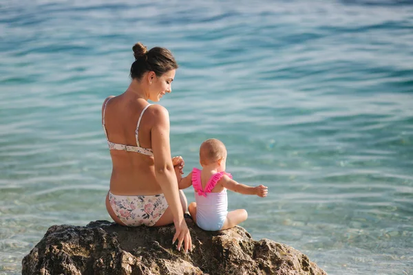 Back view of happy mom with little baby girl sits on stone in front of sea — Stock Photo, Image