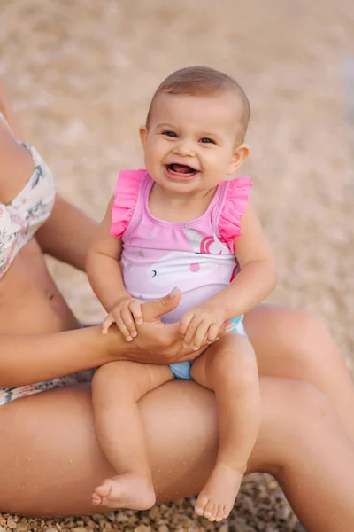 Fetiţa stă în genunchi pe plaja a treia. Tânăra mamă frumoasă cu copilul ei drăguț. Închiderea zâmbetului bebelușului — Fotografie, imagine de stoc