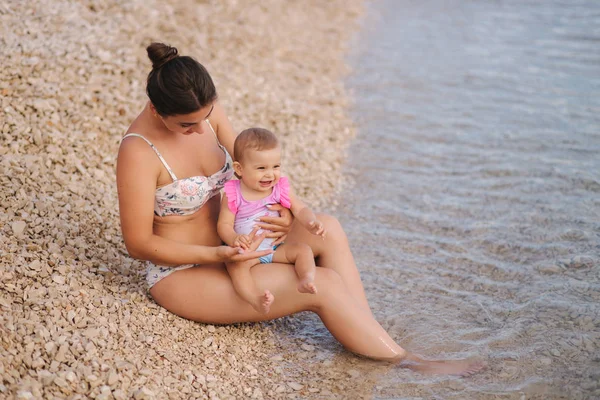 Tânăra mamă se joacă cu fetița ei pe plajă lângă mare. Mama si fiica in costum de baie — Fotografie, imagine de stoc