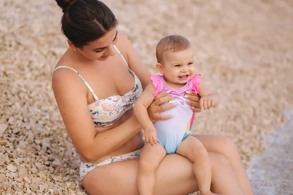 Tânăra mamă se joacă cu fetița ei pe plajă lângă mare. Mama si fiica in costum de baie — Fotografie, imagine de stoc