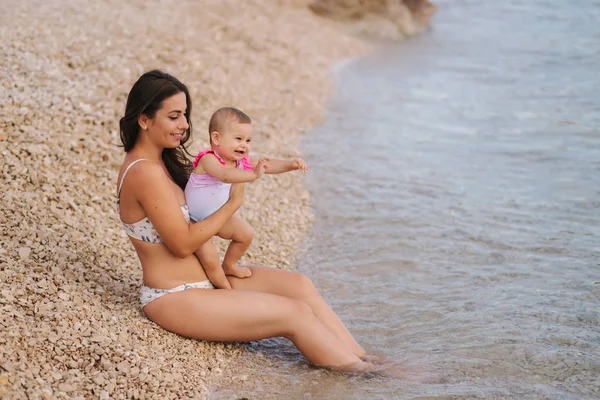 Frumoasă mamă tânără cu fetița ei drăguță care se bucură de soarele de vară pe plajă. Fetele fericite în costum de baie lângă mare. Copil râzând — Fotografie, imagine de stoc