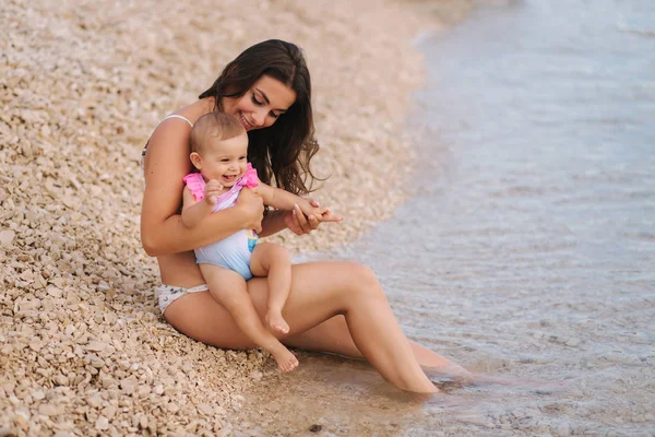 Mooie jonge moeder met haar schattige baby meisje genieten van zomer zon op het strand. Gelukkige meisjes in zwempak aan zee. Baby lachen — Stockfoto