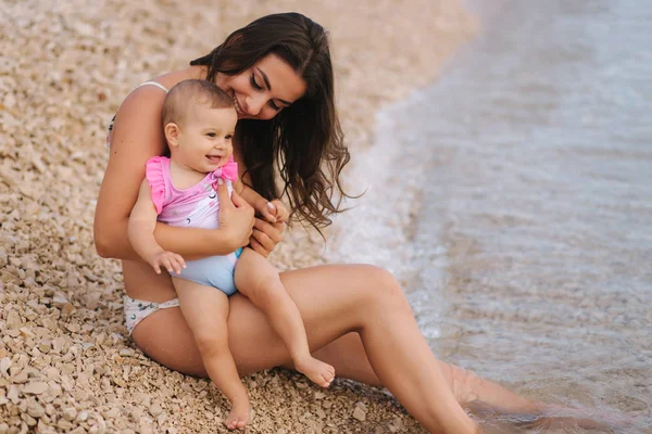 Frumoasă mamă tânără cu fetița ei drăguță care se bucură de soarele de vară pe plajă. Fetele fericite în costum de baie lângă mare. Copil râzând — Fotografie, imagine de stoc