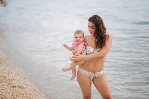 Mama ține fetița drăguță pe mâini pe plaja din mare. Mama si fiica in costum de baie — Fotografie, imagine de stoc