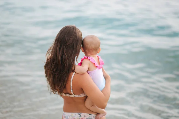 Vedere din spate a mamei țineți fetița pe mâini pe plaja din mare. Mama si fiica in costum de baie — Fotografie, imagine de stoc