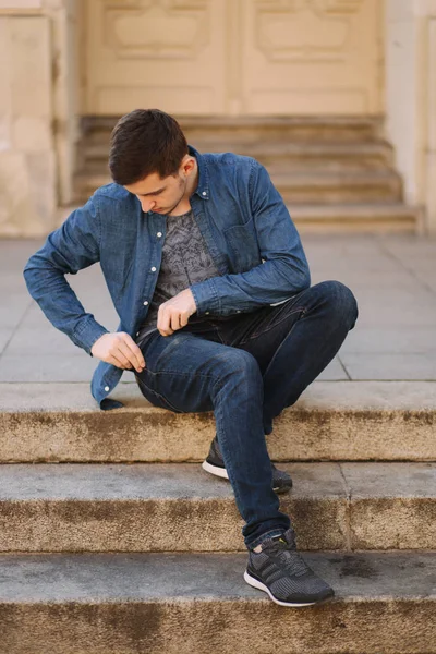 Hombre guapo sitiing en las escaleras cerca del parque y poner la mano en un bolsillo para sacar la cubierta para auriculares inalámbricos — Foto de Stock