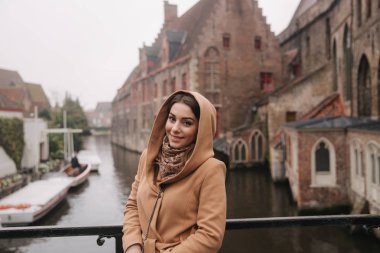 Bealgium, Brugge köprüsünde kahverengi ceket ayakta güzel kadın portresi. Kadın bir Hood koymak