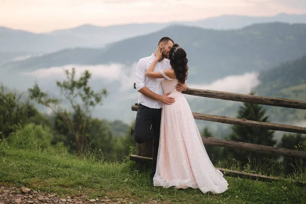 Bájos menyasszony ölelés neki jóképű vőlegény a hegyekben. Gyönyörű esküvő pár séta. Zöld gackground — Stock Fotó