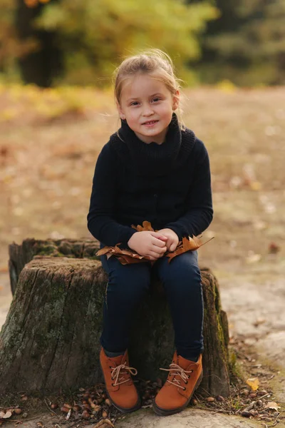 Uma menina em roupas pretas está sentada em um toco e brincando com folhas de outono. Cabelo loiro menina — Fotografia de Stock