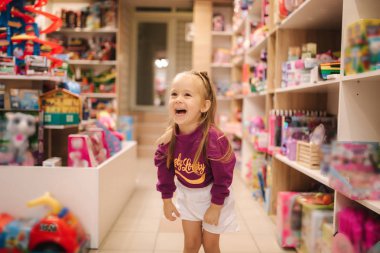 Adorable little girl shopping for toys. Cute female in toy store. Happy young girl selecting toy clipart