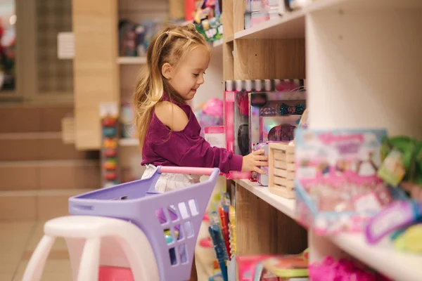 Niña con carrito de compras pequeño en el centro comercial para niños. Chica feliz elegir qué comprar en la tienda de juguetes — Foto de Stock