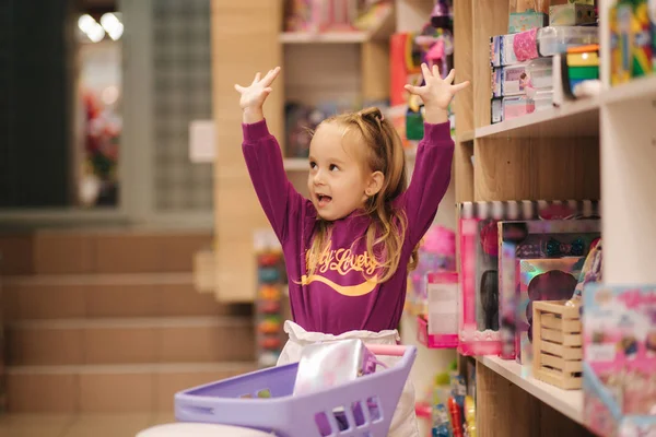 Маленькая девочка с маленькой тележкой в детском торговом центре. Счастливая девушка выбирает, что купить в магазине игрушек — стоковое фото