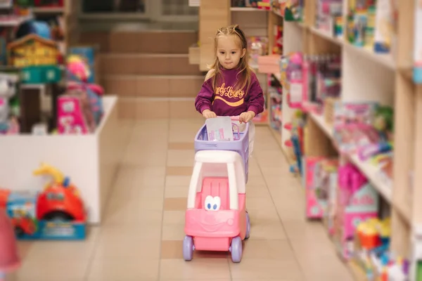 Маленькая девочка с маленькой тележкой в детском торговом центре. Счастливая девушка выбирает, что купить в магазине игрушек — стоковое фото
