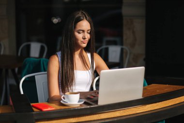 Young business lady work on laptop and drinking a coffee. Beautiful girl sitting in cafe outside. Woman using phone