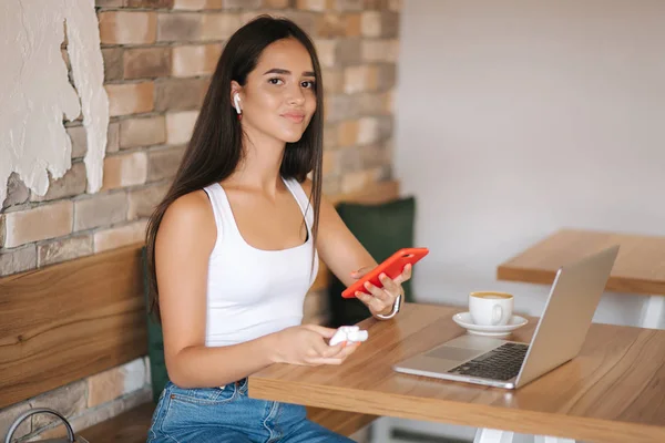 有吸引力的年轻女孩使用无线耳机和笔记本电脑工作。穿白色T恤和牛仔布的女人。学生在咖啡馆学习 — 图库照片