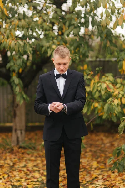 Schöner Bräutigam im Hochzeitsanzug draußen, Herbst. Hintergrund des gelben Baumes. Mann im schwarzen Anzug mit schöner Krawattenfliege — Stockfoto