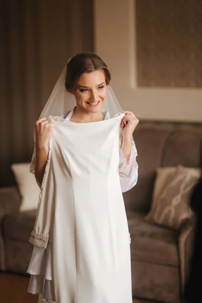 Retrato de bela noiva em pé junto à janela em casa e experimentando o vestido de noiva. Noiva encantadora com vestido de noiva branco — Fotografia de Stock