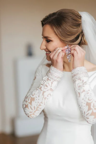 Κοντινό πορτρέτο της όμορφης νύφης που στέκεται δίπλα στο παράθυρο στο σπίτι και δένει σκουλαρίκια. Γοητευτική νύφη με λευκό νυφικό — Φωτογραφία Αρχείου