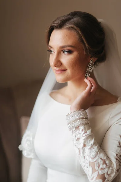 Close up retrato de bela noiva em pé junto à janela em casa e amarrando em brincos. Noiva encantadora em vestido de casamento branco — Fotografia de Stock