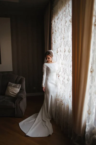 Schöne Braut, die zu Hause am Fenster steht. Charmante Braut im weißen Hochzeitskleid — Stockfoto