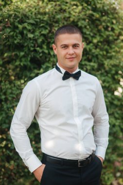 Siyah papyonlu beyaz gömlekli genç adam dışarıda duruyor.