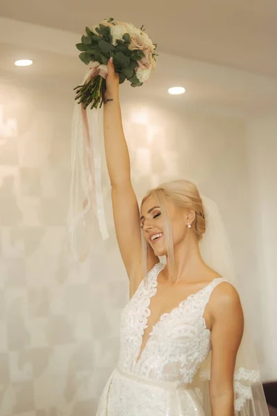 Элегантная невеста в красивом свадебном платье с букетом дома. Блондинка с красивым макияжем и прической — стоковое фото