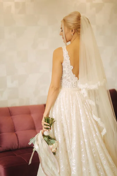 Elegantní nevěsta v krásných svatebních šatech s kyticí doma. Blond vlasy žena s krásný make-up a účes — Stock fotografie