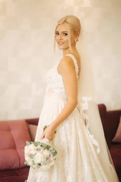 Novia elegante en hermoso vestido de novia con ramo en casa. Mujer de cabello rubio con hermoso maquillaje y peinado — Foto de Stock