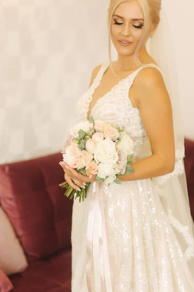 Noiva elegante em belo vestido de noiva com buquê em casa. Mulher de cabelo loiro com bela maquiagem e penteado — Fotografia de Stock