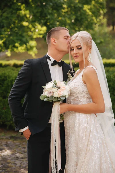 Guapo novio abrazar a su hermosa novia. Recién casados caminando por el parque. Mujer pelo rubio en vestido de novia elegante — Foto de Stock