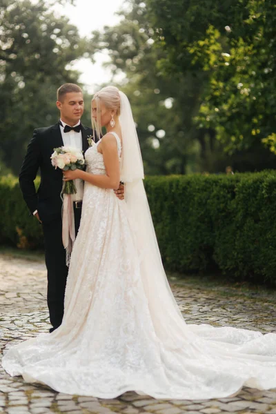 Bonito noivo abraçar sua linda noiva. Recém-casados a passear no parque. Mulher de cabelo loiro em vestido de noiva elegante — Fotografia de Stock