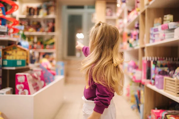 Vista posterior de adorable niña comprando juguetes. Linda hembra en la tienda de juguetes. Feliz chica joven seleccionando juguete — Foto de Stock